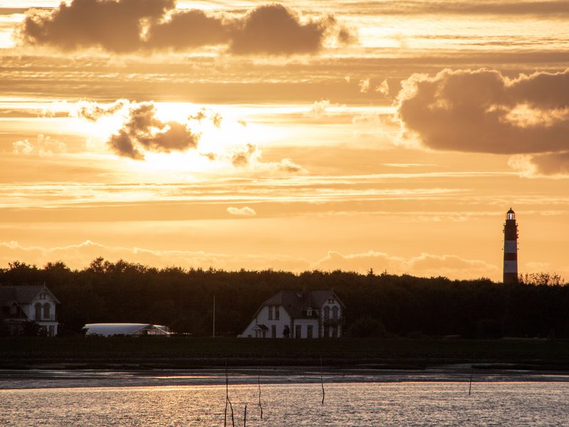 Der rot-weiß geringelte Leuchtturm von Wittdün auf Amrum im Gegenlicht der untergehenden Sonne, davor Häuser hinter dem Deich