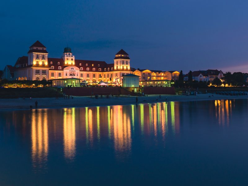 Das beleuchtete Kurhaus Binz zur blauen Stunde, davor der Strand und die Ostsee, in der sich die Lichter der Promenade spiegeln