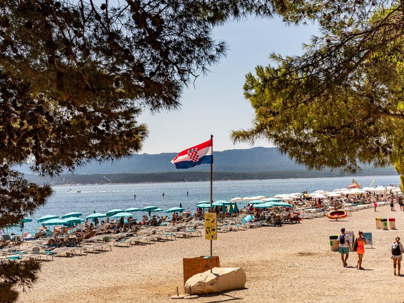 Der Kiesstrand Zlatni Rat bei Bol auf Brač: kroatische Flagge über Liegestuhlreihen im Schatten der Kiefern, dahinter das Meer und die Berge von Hvar