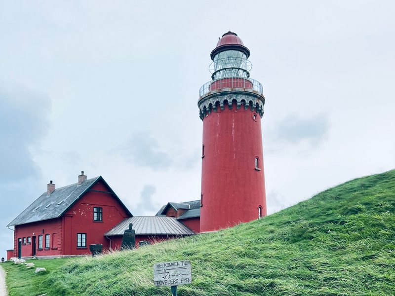Der rote Leuchtturm Bovbjerg Fyr über der jütländischen Westküste, daneben das rote Leuchtturmwärterhaus und ein Wiesenhang mit dem Schild „Velkommen til Bovbjerg Fyr"