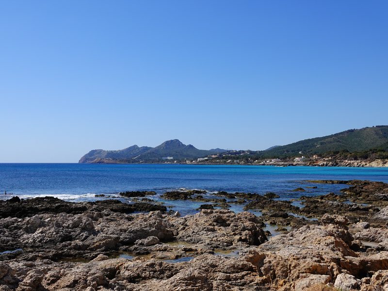Die Felsküste von Cala Rajada im Nordosten Mallorcas, dahinter eine türkise Bucht und die bewaldeten Berge der Serra de Llevant
