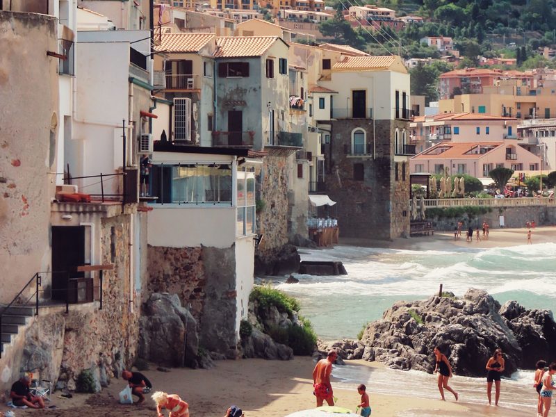Die Altstadthäuser von Cefalù stehen direkt über dem Wasser, rechts der Stadtstrand mit Sonnenschirmen, dahinter steigt der bewaldete Hang an