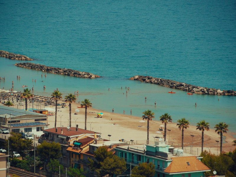 Der Adriastrand von Cupra Marittima mit Palmenreihe, Liegestühlen und vorgelagerten Steinbuhnen, dahinter die Häuser des Ortes — zeigt nicht den genannten Ort