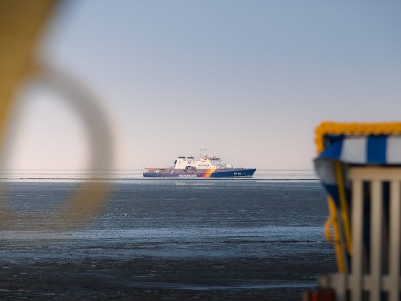 Ein Schiff der Bundespolizei-Küstenwache fährt durchs Wattenmeer vor Cuxhaven-Duhnen, im Vordergrund unscharf ein Rettungsring und ein Strandkorb