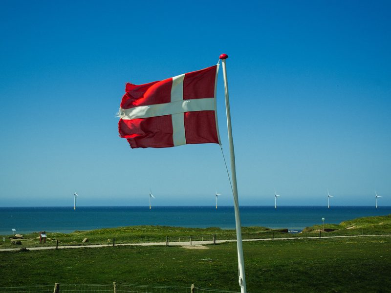 Eine wehende dänische Flagge auf einer Küstenwiese, dahinter das Meer mit einem Offshore-Windpark am Horizont — zeigt nicht den genannten Ort