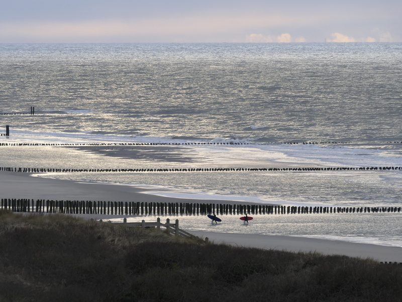 Zwei Surfer tragen ihre Bretter über den Strand von Domburg, davor Reihen von Buhnenpfählen im flachen Wasser der Nordsee