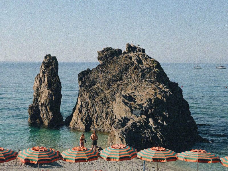 Reihen rot-grün gestreifter Sonnenschirme am Strand von Monterosso al Mare, dahinter der markante Felsen im flachen Wasser — zeigt nicht den genannten Ort