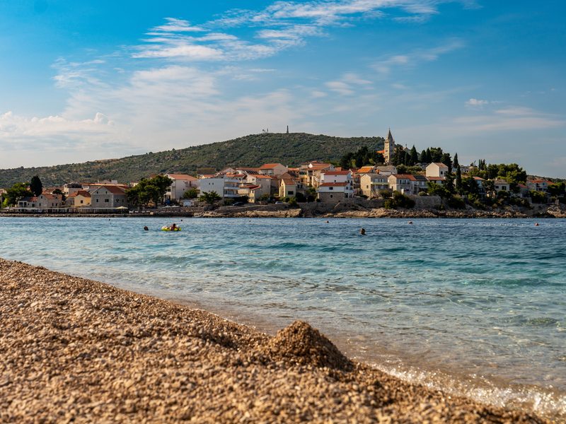 Blick über eine Kiesbucht auf die Altstadt von Primošten, die sich mit ihrem Kirchturm über eine bewaldete Halbinsel zieht — zeigt nicht den genannten Ort