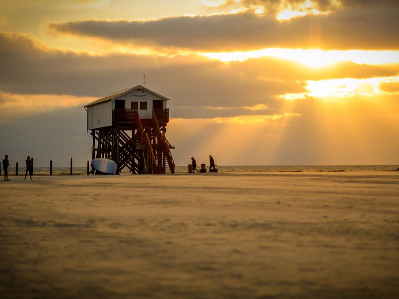 Ein Pfahlbau am Strand von Sankt Peter-Ording im Gegenlicht der untergehenden Sonne, davor spielen Kinder im Sand