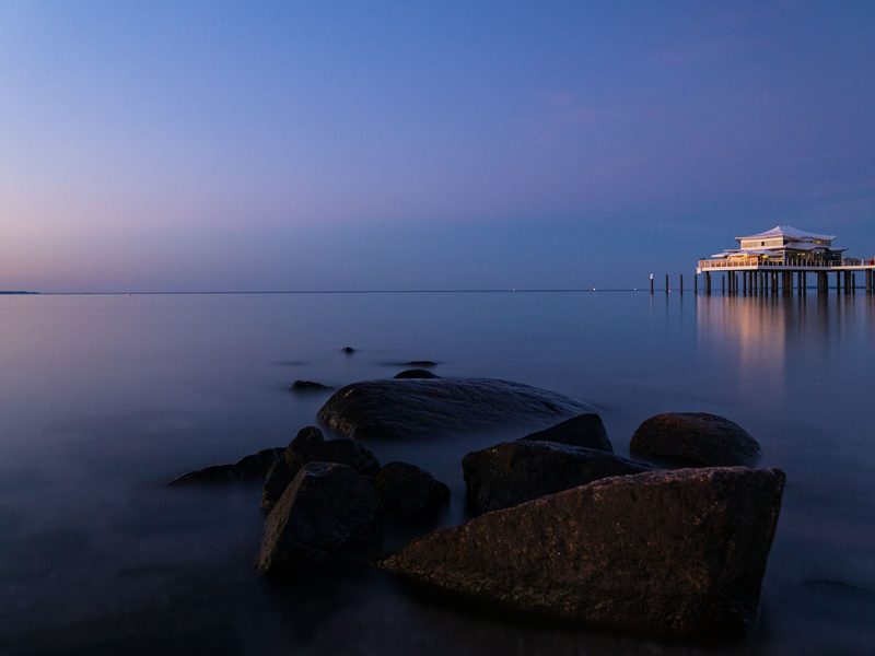 Das Teehaus auf der Seebrücke von Timmendorfer Strand zur blauen Stunde, davor Findlinge im spiegelglatten Wasser der Ostsee