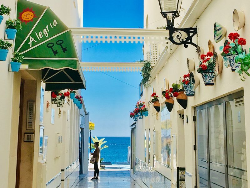 Weiß getünchte mediterrane Gasse mit blühenden Wandtöpfen und Blick auf das Meer — zeigt nicht den genannten Ort