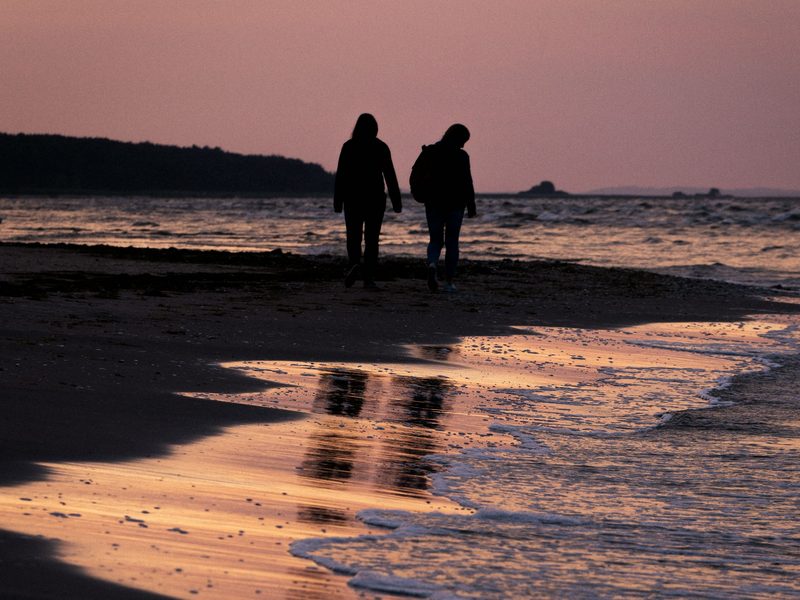 Zwei Menschen gehen bei Sonnenuntergang am Strand von Usedom entlang, ihre Silhouetten spiegeln sich im nassen Sand