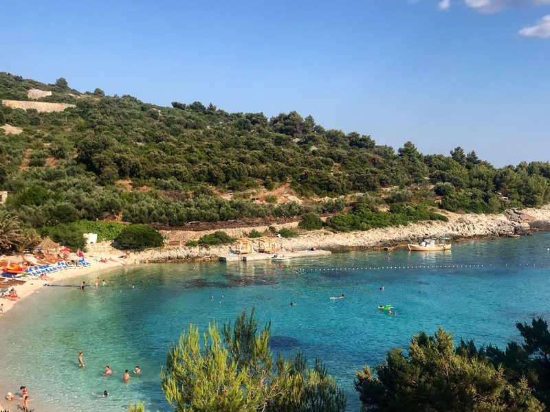 Eine Badebucht auf der Insel Vis mit hellem Kiesstrand und türkisem Wasser, dahinter ein Hang aus Kiefern und Olivenbäumen — zeigt nicht den genannten Ort