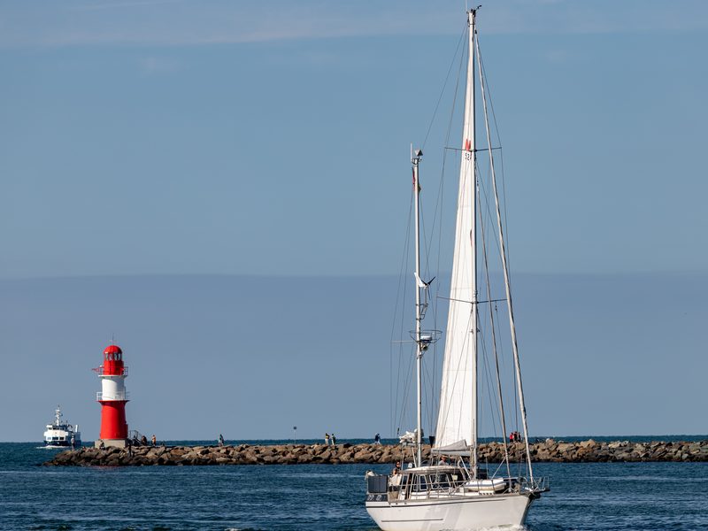 Das rot-weiße Mole-Leuchtfeuer an der Warnemünder Hafeneinfahrt, davor kreuzt ein Segelboot mit gesetztem Großsegel